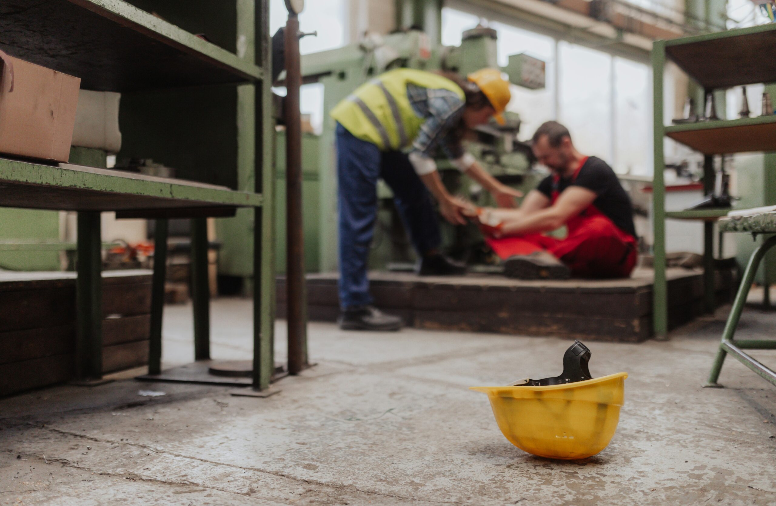 A yellow safety helmet lies on a concrete factory floor in the foreground, slightly scuffed and tipped on its side, while in the blurred background two industrial workers are inside a workshop with heavy machinery, one seated on the ground clutching his leg and wearing red overalls, the other standing nearby in a reflective safety vest and hard hat, bending down to assist him, suggesting a workplace accident or injury in an industrial manufacturing environment.