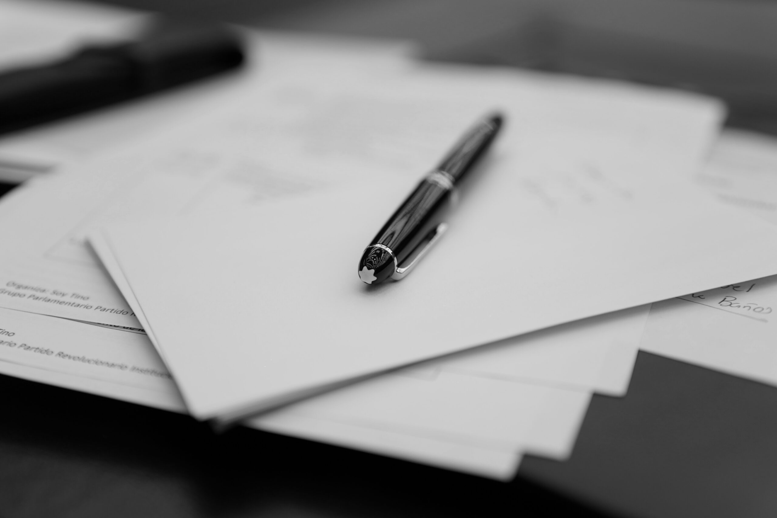 A pen rests on top of a sheet of paper on a wooden desk, creating a simple workspace scene.