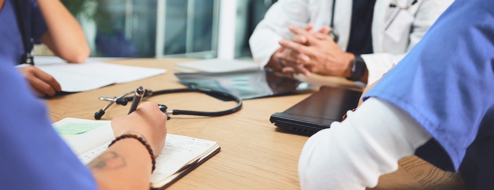 Close-up of medical staff around a table with a stethoscope, notebook, and laptop during a discussion.