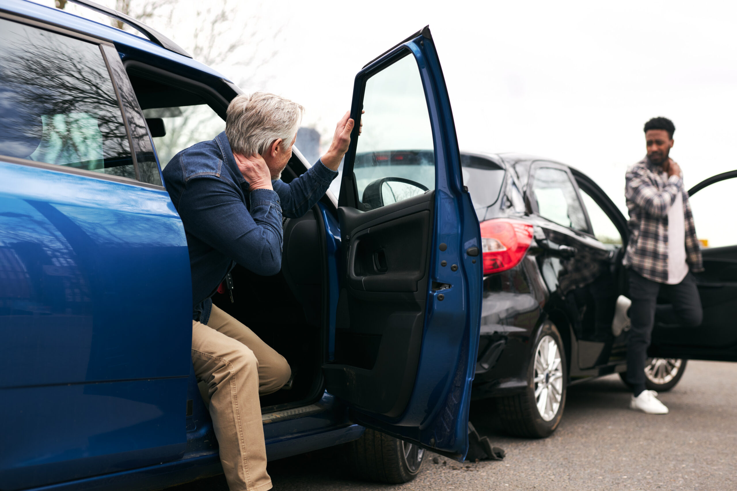 A man exits his damaged blue car holding his head while another man stands by a dented black car after a crash.