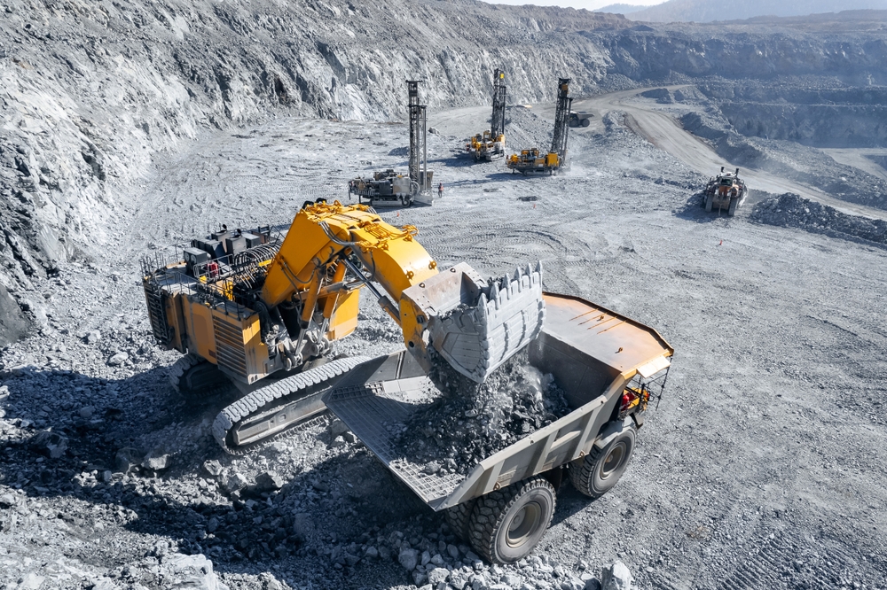 An aerial view of a large excavator loading rock into a mining dump truck at an open-pit quarry.