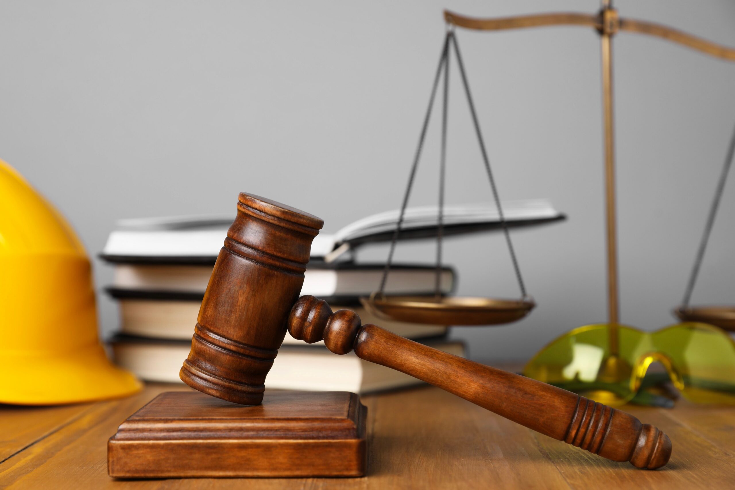 Wooden gavel beside a yellow hard hat, safety glasses, law books, and scales of justice on a desk