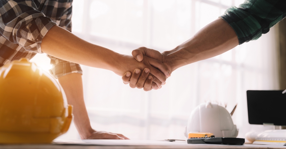 Two people shaking hands over a desk with hard hats, blueprints, and a calculator